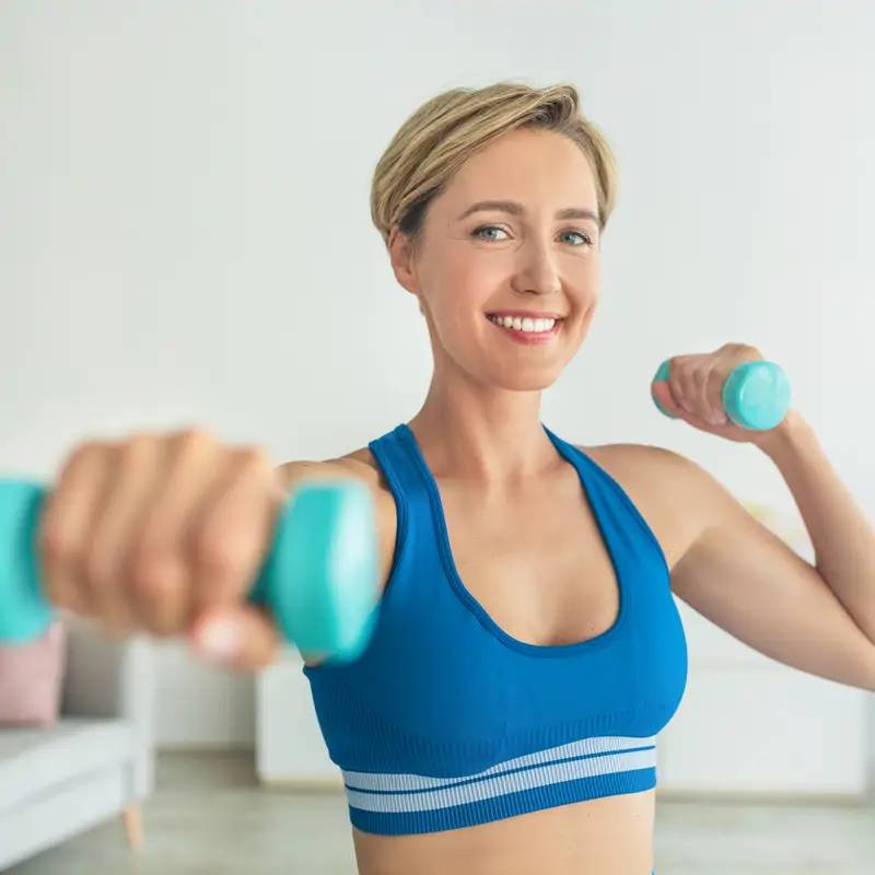 Mujer sonriente de mediana edad haciendo ejercicio con dos mancuernas en casa